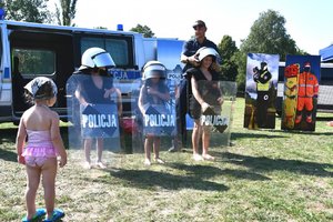 troje dzieci stoi z policyjnymi tarczami, z tyłu policjant zakładający hełm jednemu z nich, na pierwszym planie mała dziewczynka zwrócona w stronę dzieci