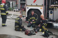 zdjęcie przedstawia trzech siedzących na ulicy strażaków zmęczonych po akcji gaśniczej