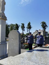 Na fotografii widać cmentarz. Przed jednym z pomników stoją dwaj policjanci i oddają honor.