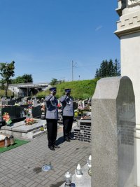 Na fotografii widać cmentarz. Przed jednym z pomników stoją dwaj policjanci i oddają honor.