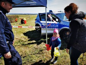 dzielnicowy rozmawiaz małą dziewczynką oraz jej mamą, w tle policyjny radiowóz