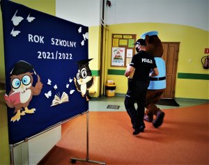 od lewej szkolna dekoracja &quot;Rok szkolny 2021/2022&quot;, dalej policjant w mundurze prowadzi maskotkę sierżanta Hektora, scena ma miejsce na szkolnym korytarzu