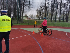 policjant przygląda się jeżdżącemu na rowerze chłopcu, scena ma miejsce na szkolnym boisku